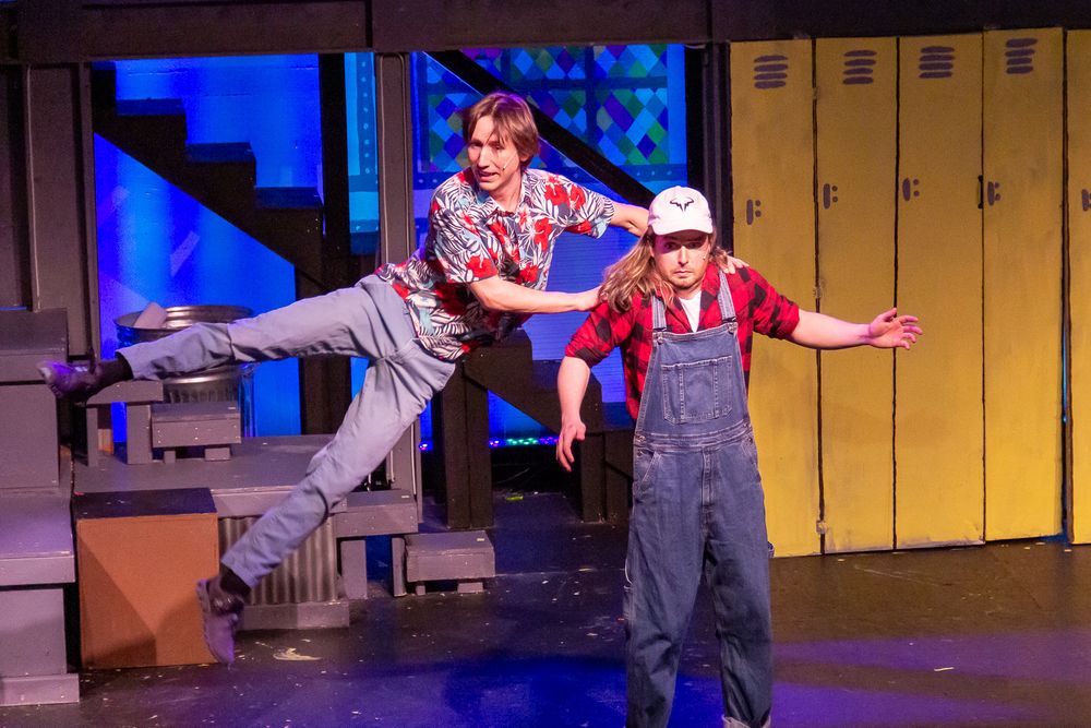 Two performers in overalls dance energetically on a stage with lockers in the background.