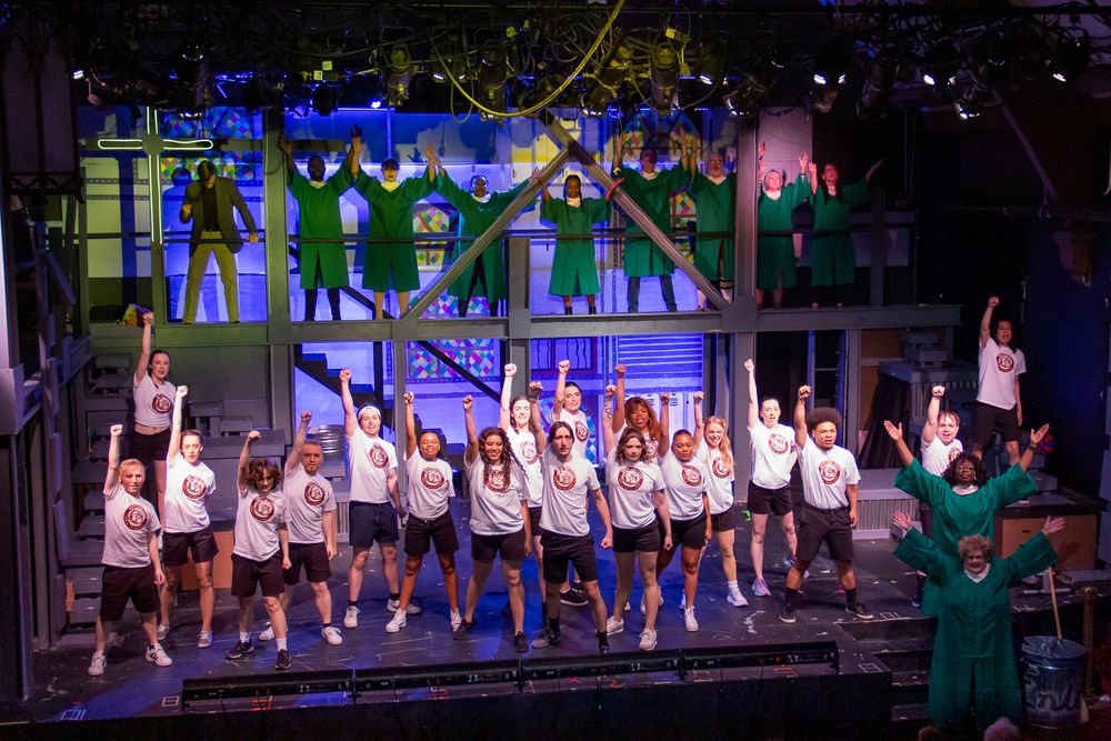 Cast members perform a synchronized dance number on stage wearing matching white outfits.