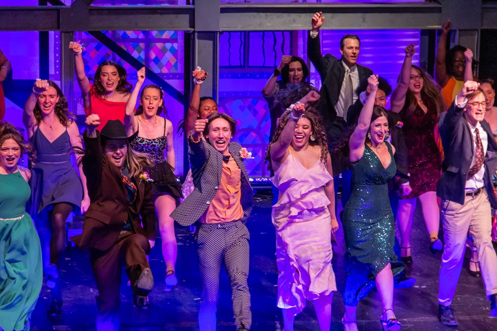 Musical theater performers dancing in colorful costumes under purple stage lighting.
