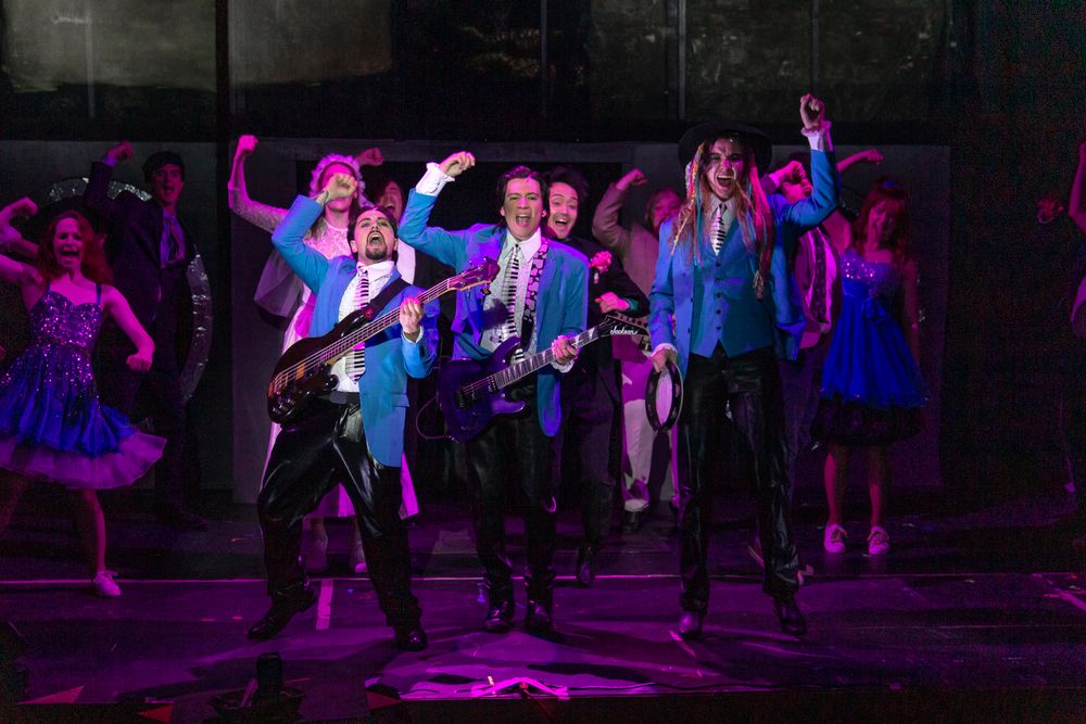 Performers in dark costumes execute a dance routine under dramatic purple stage lighting.