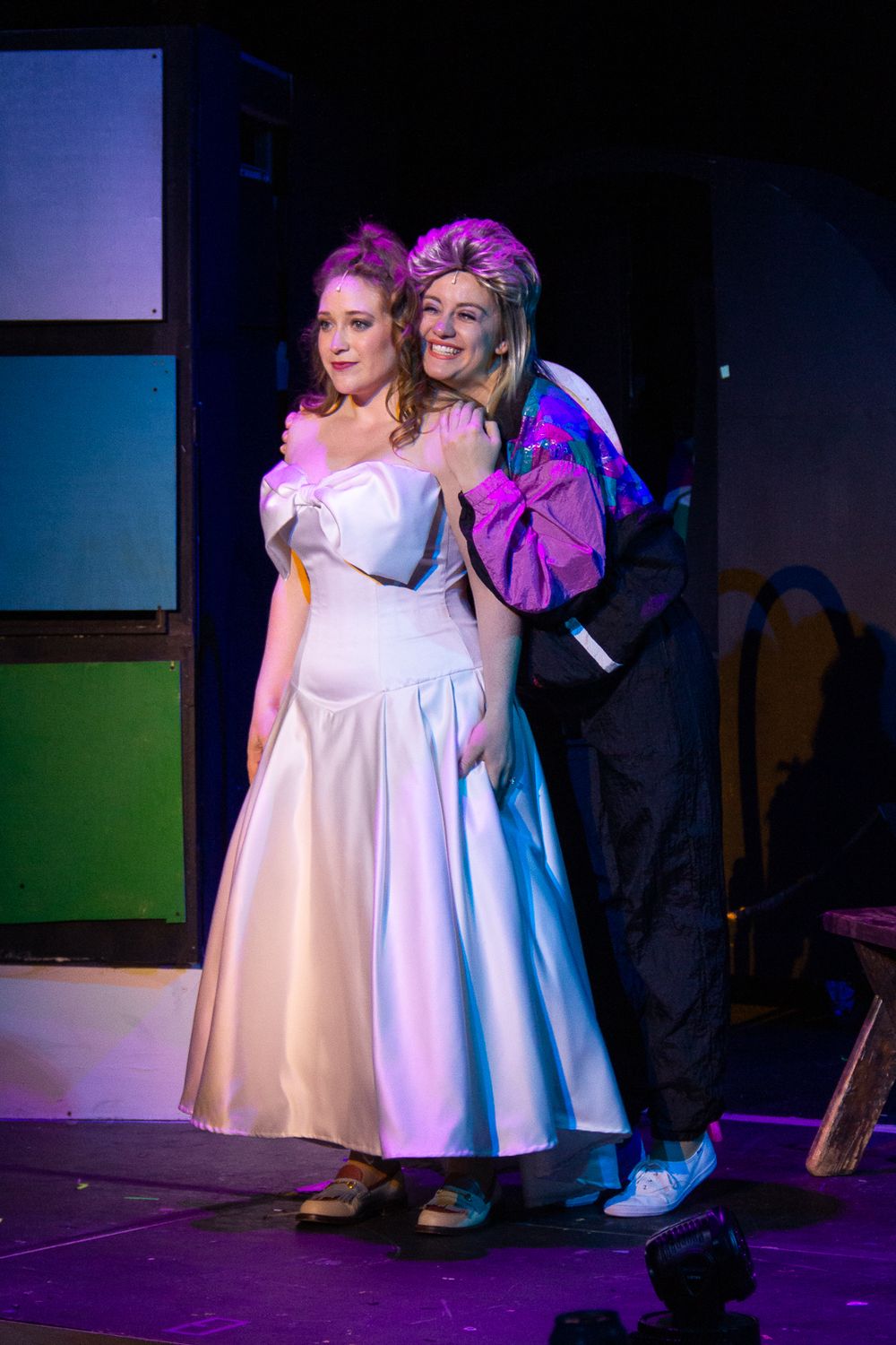 Two performers in contrasting outfits pose together during a stage production with purple lighting.