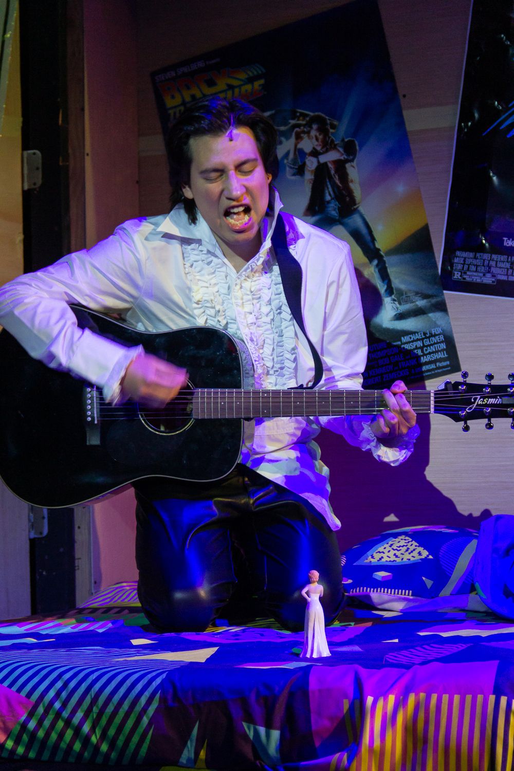 Performer in white outfit plays acoustic guitar during theatrical performance with dramatic blue lighting.