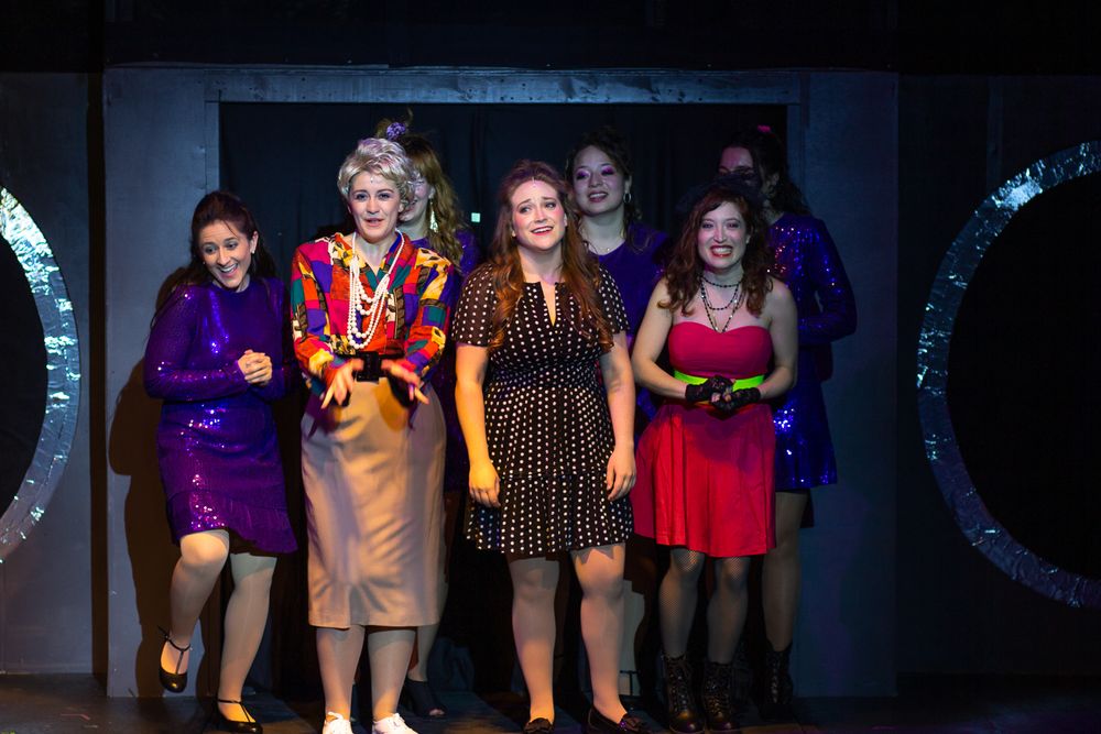 Cast members in colorful formal attire perform on a theatrical stage set.