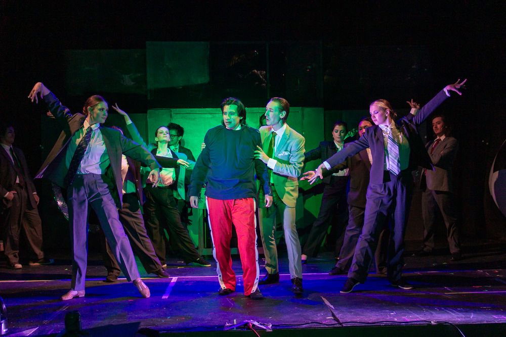 Performers dance energetically on stage with dramatic green and blue lighting during a theatrical production.