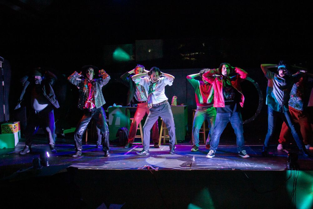 Dancers perform a synchronized routine on a dark stage illuminated by colorful spotlights.