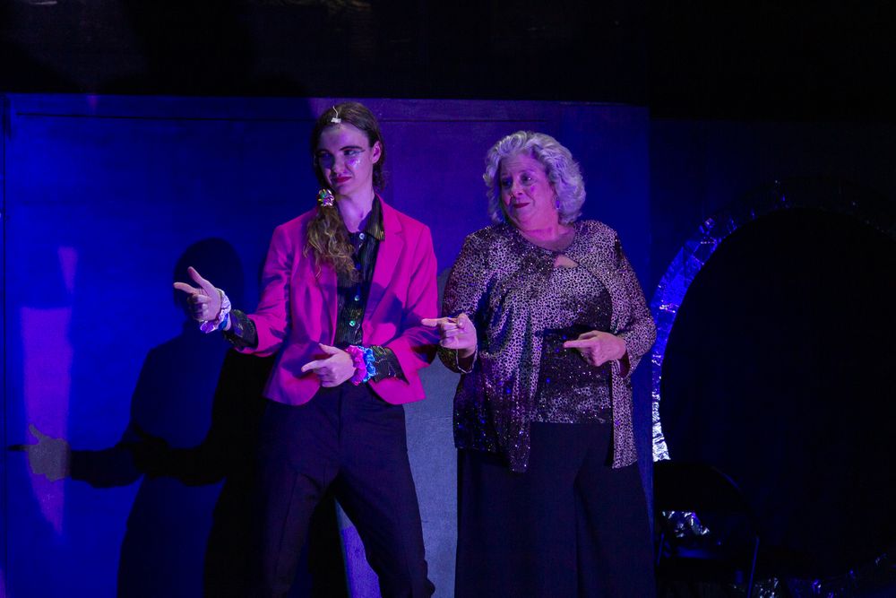 Two performers on stage bathed in dramatic purple lighting during a theatrical scene.