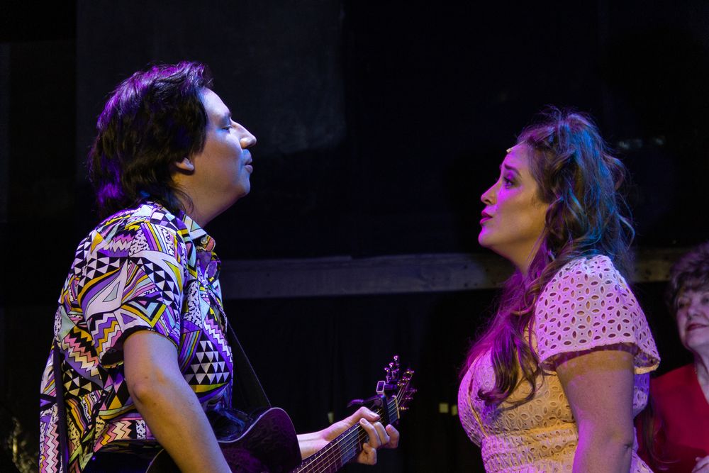 Two performers share an intimate moment during a musical theater scene under pink stage lighting.