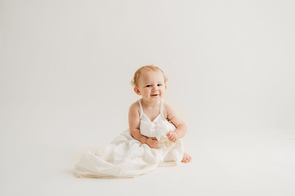 Baby wearing white dress sitting on floor during minimalist portrait session.