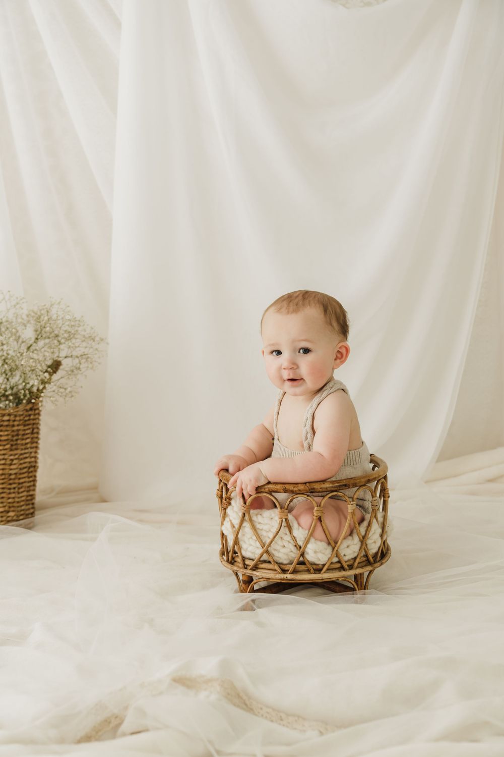 Baby in beige romper sitting in decorative basket against white studio backdrop.