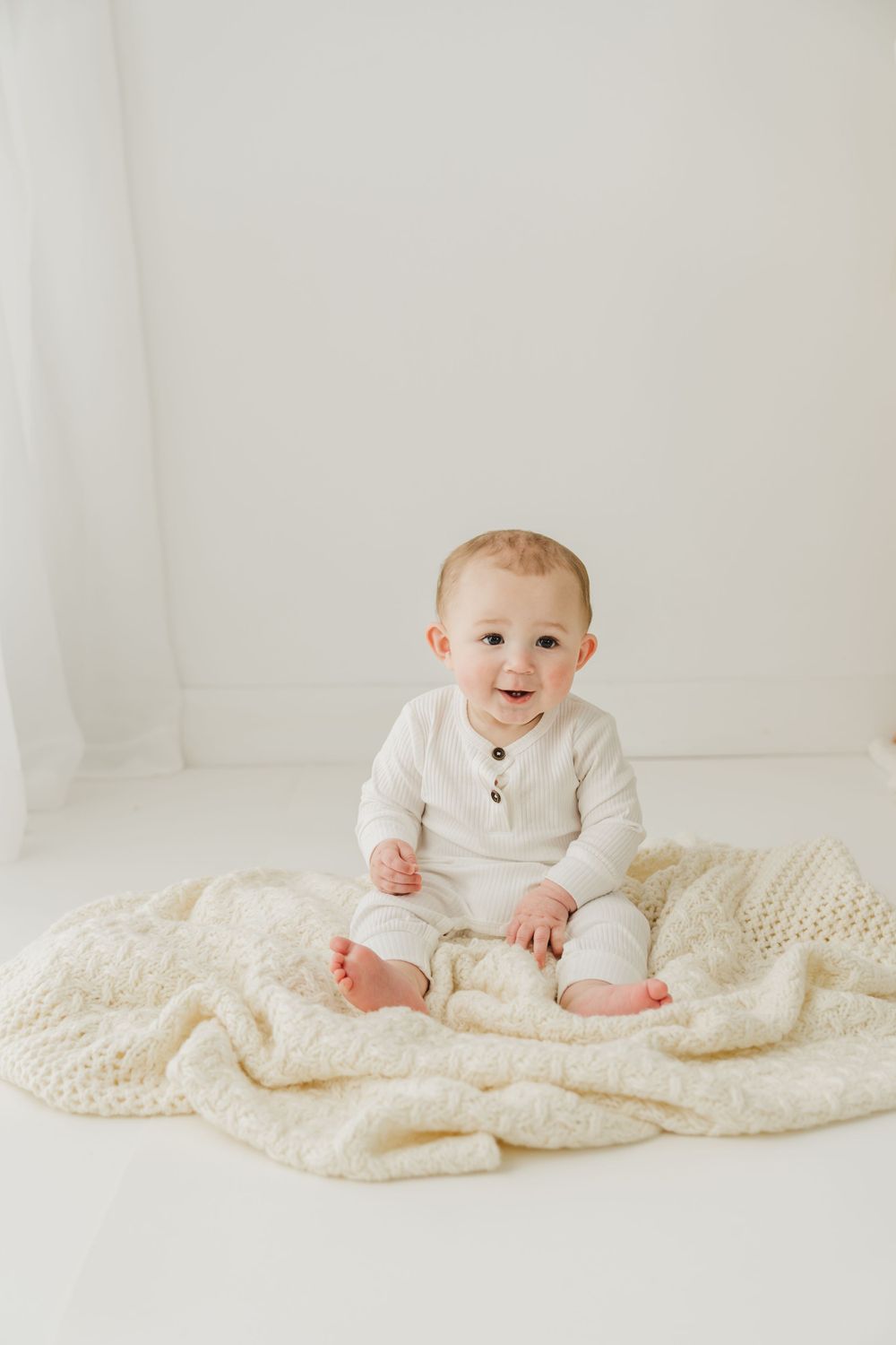 Baby in white henley outfit smiling on knitted blanket during portrait session.