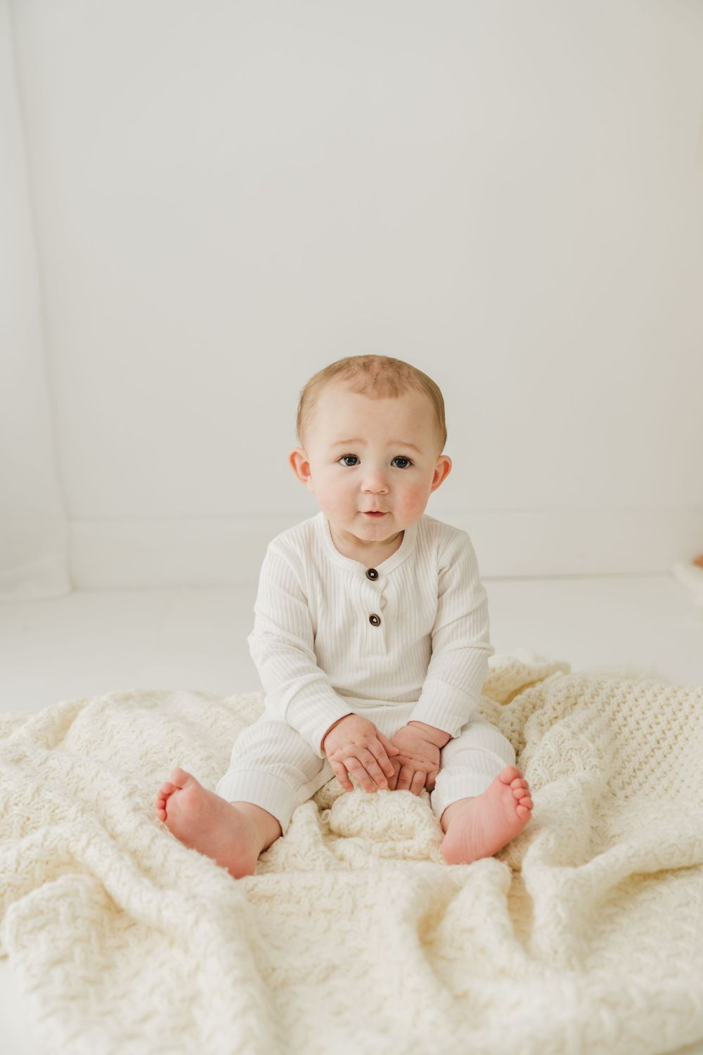 Baby in white knit outfit sitting on cream blanket during studio photography session.