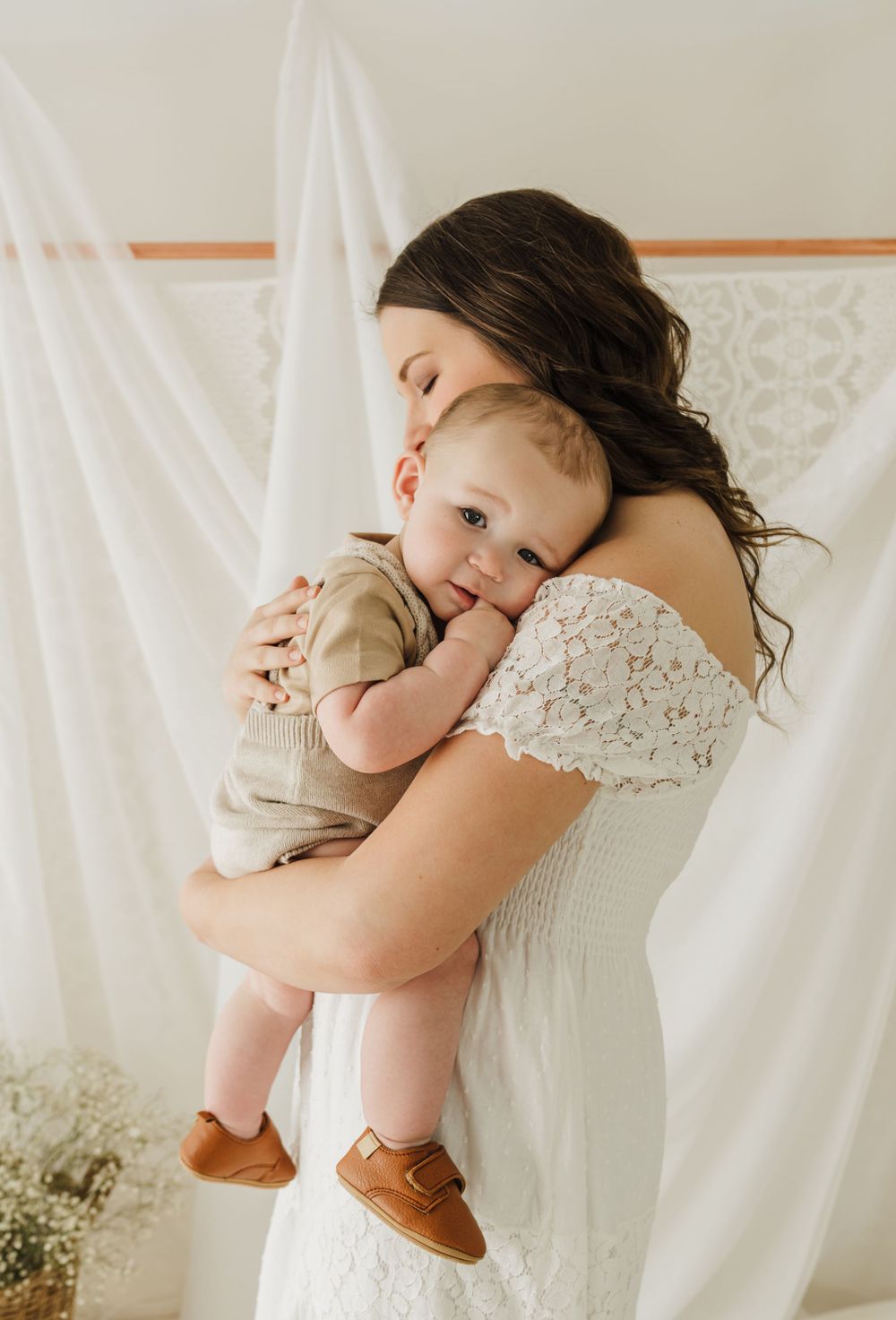 A tender moment between a mother in a white lace dress embracing her baby wearing brown shoes.