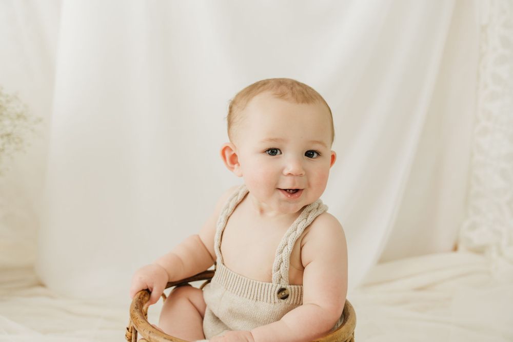 Baby sitting in wooden basket wearing beige romper during studio photoshoot.