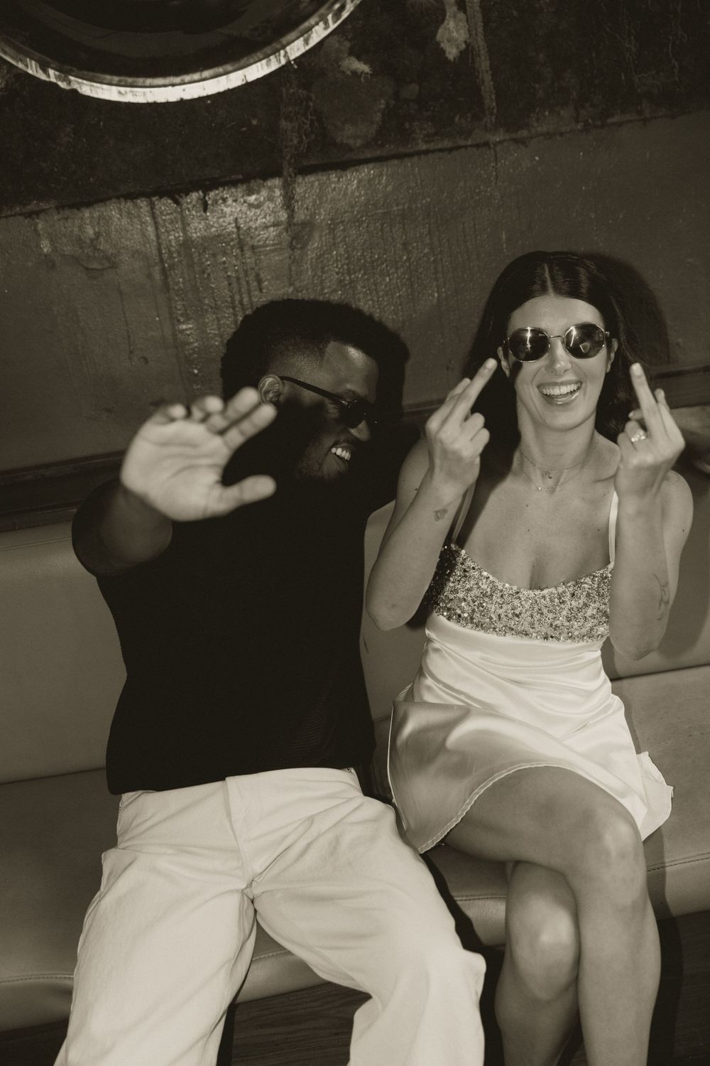 Two friends in sunglasses pose playfully in sepia-toned photo, man in black shirt and woman in sequin dress.