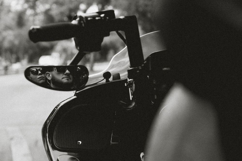 Black and white photo of a motorcycle mirror reflecting a rider wearing sunglasses on a tree-lined road.