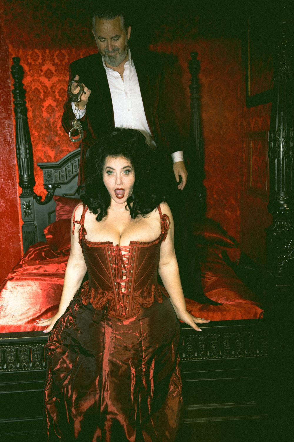 Woman in red corset gown with surprised expression stands before a man in a gothic Victorian bedroom setting.