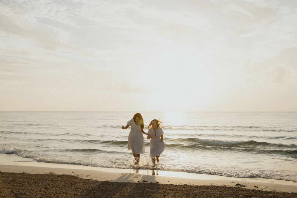 Two girls in white dresses hold hands running toward ocean waves on a sandy beach at sunset.