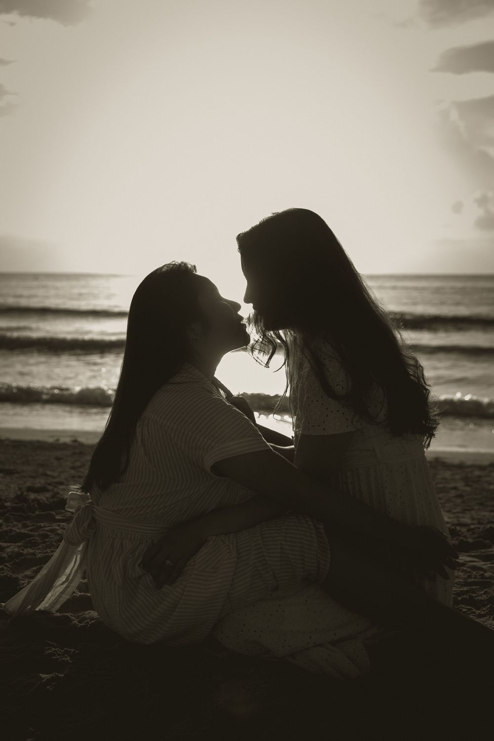Two women in white dresses share a romantic moment on a beach at sunset in a sepia-toned photo.