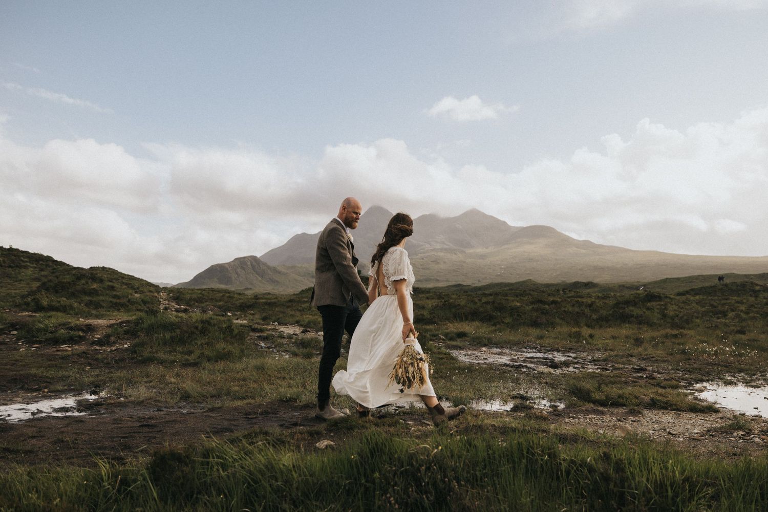 A couple walks together on the rugged Scottish highlands surrounded by mountains and mist.