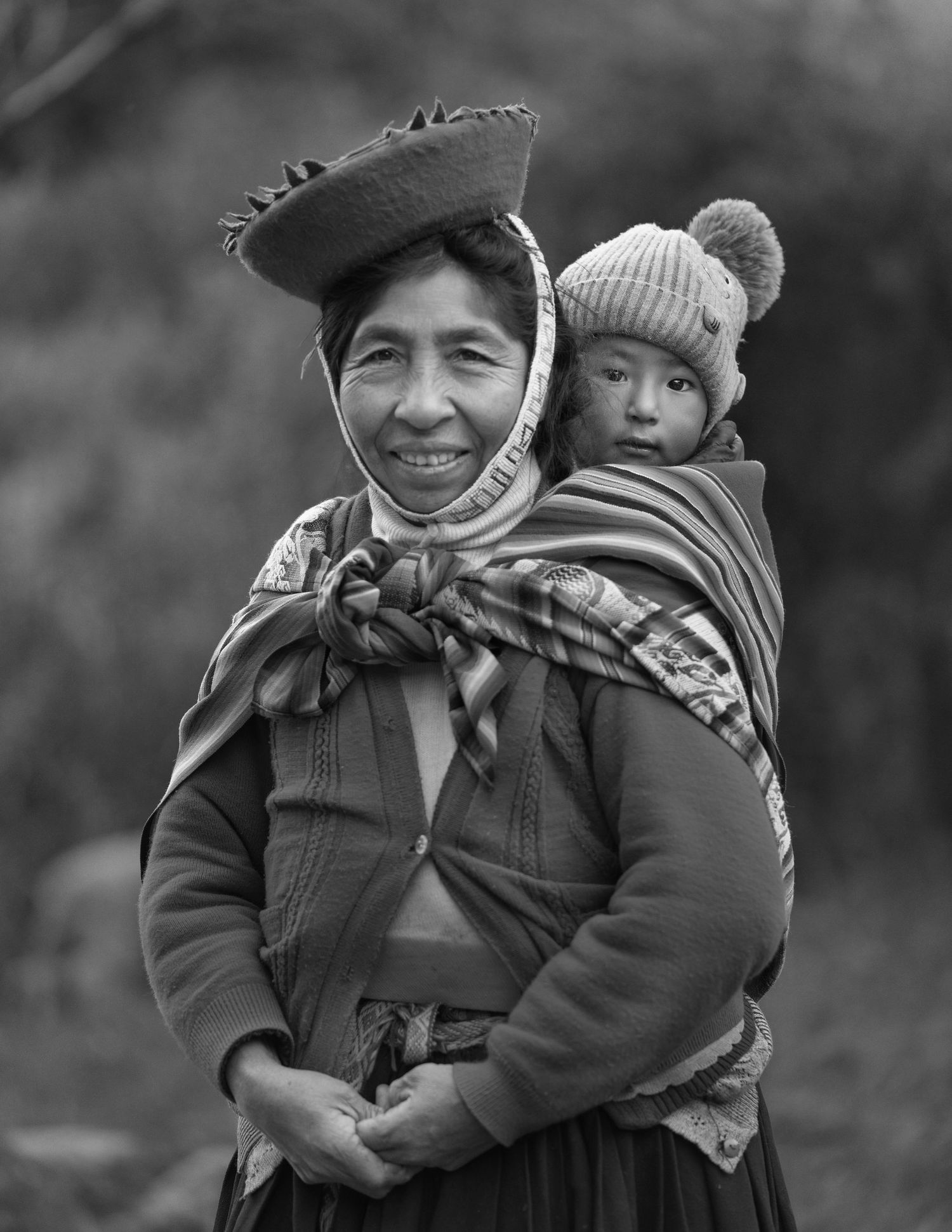 Traditional Andean woman wearing red hat and carrying child in colorful woven textile wrap.