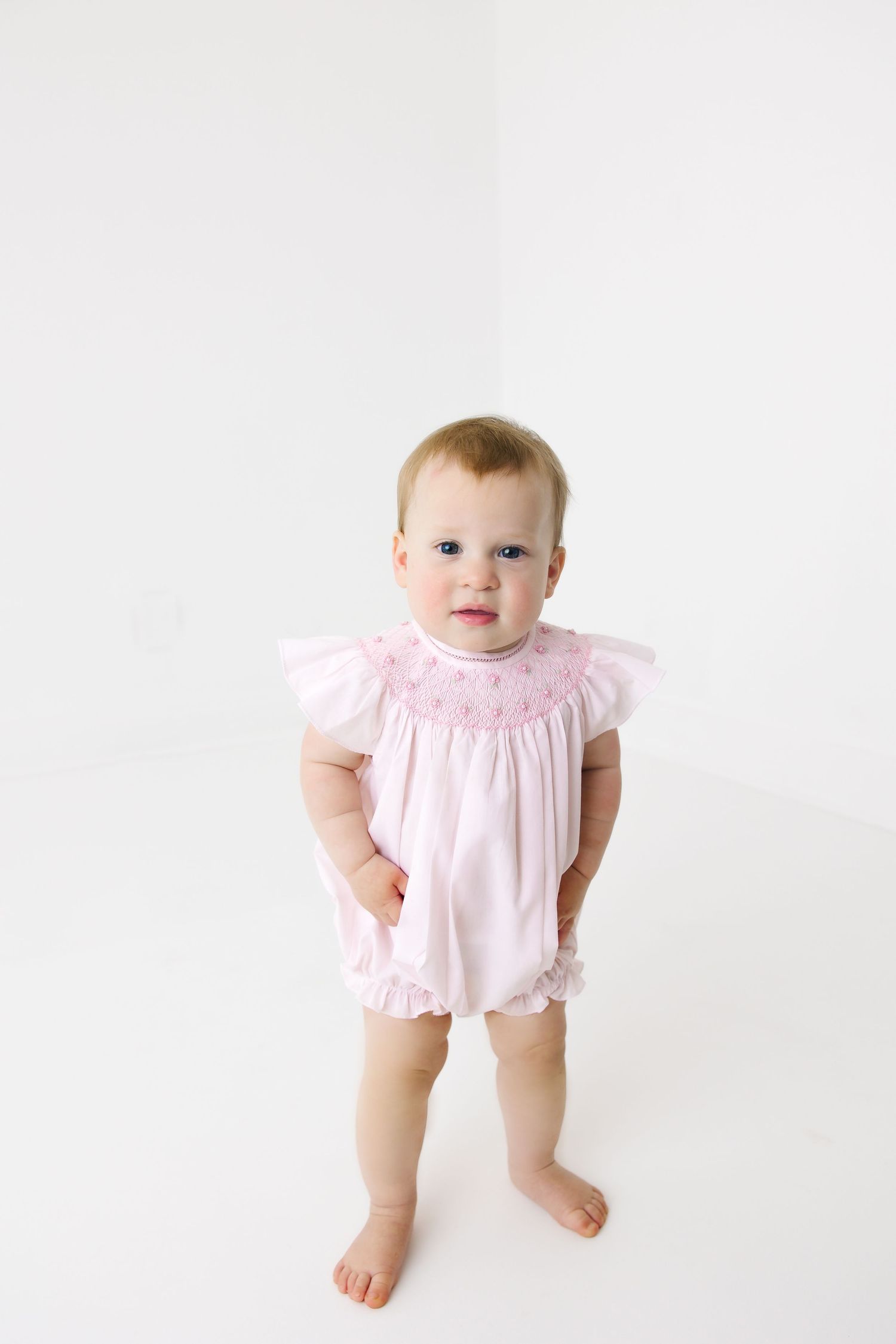 Small child standing in pink smocked romper against white background.