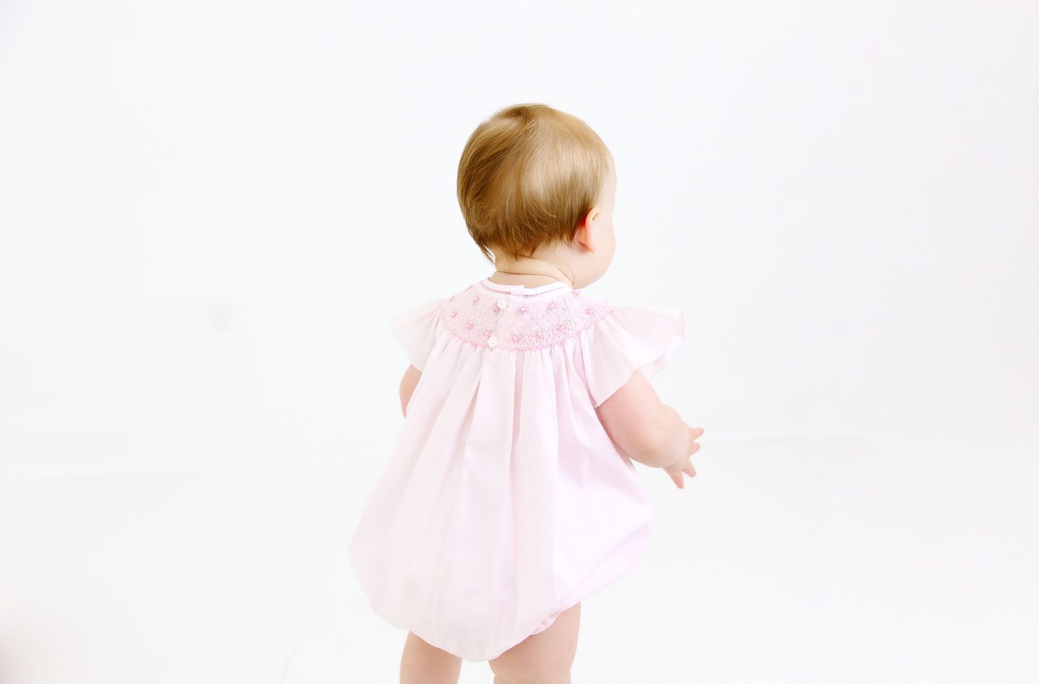 A young toddler wearing a pink dress stands facing away from the camera against a white backdrop.