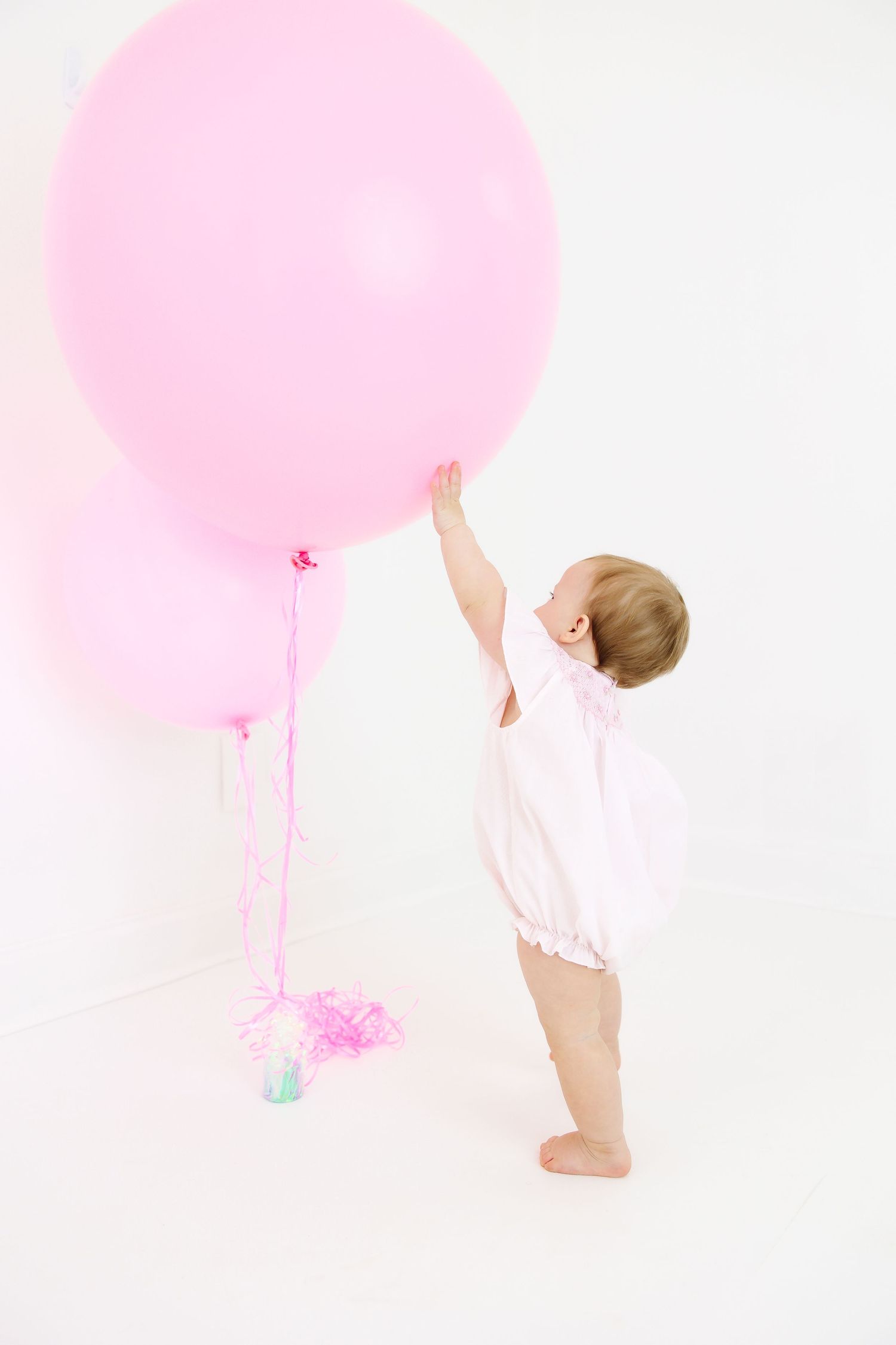 Small baby in white outfit reaching for large pink balloon in minimalist studio setting.