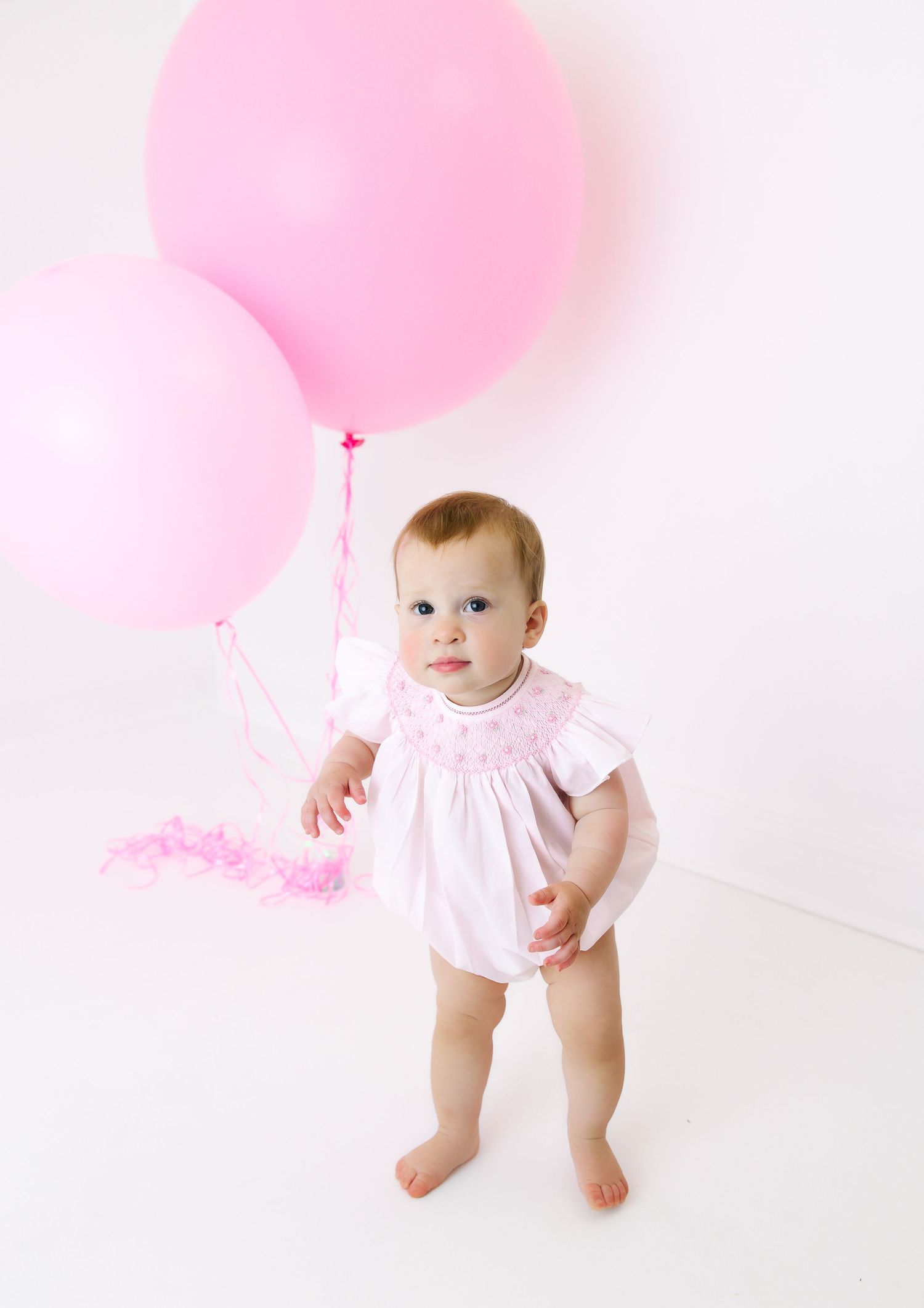 Baby in pink smock standing against white background with large pink balloons.