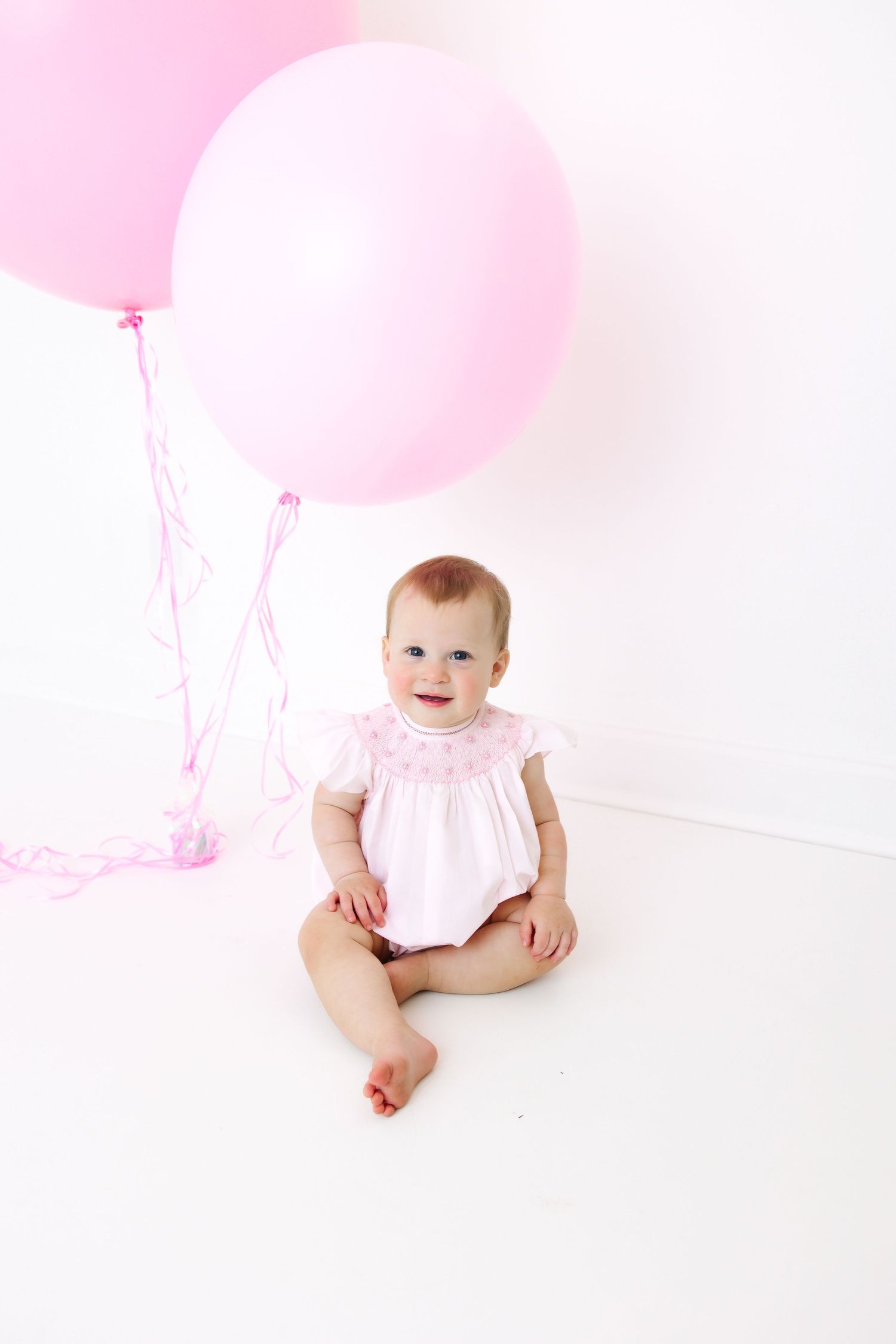 Baby in pink outfit sits on white background with pink balloons around them.