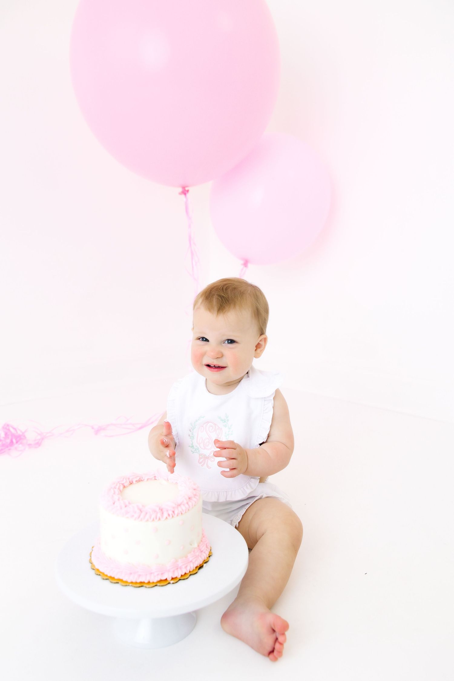 Baby with pink balloons and birthday cake sitting on white floor.