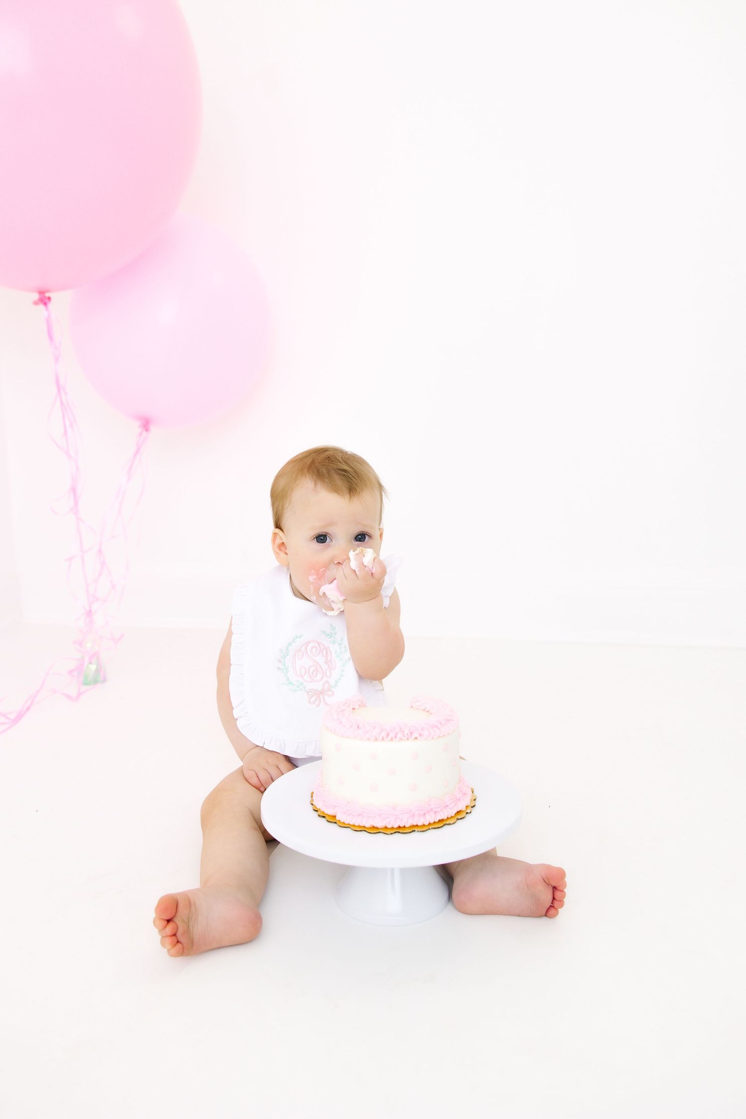 Baby in white outfit exploring pink birthday cake with balloon decoration.