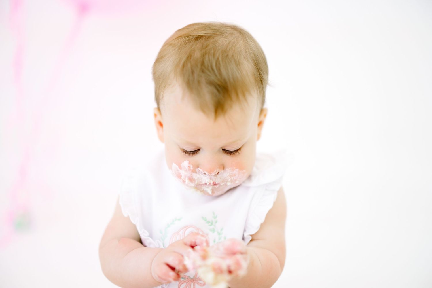 Baby in white dress reaching for pink birthday cake during first birthday photoshoot.
