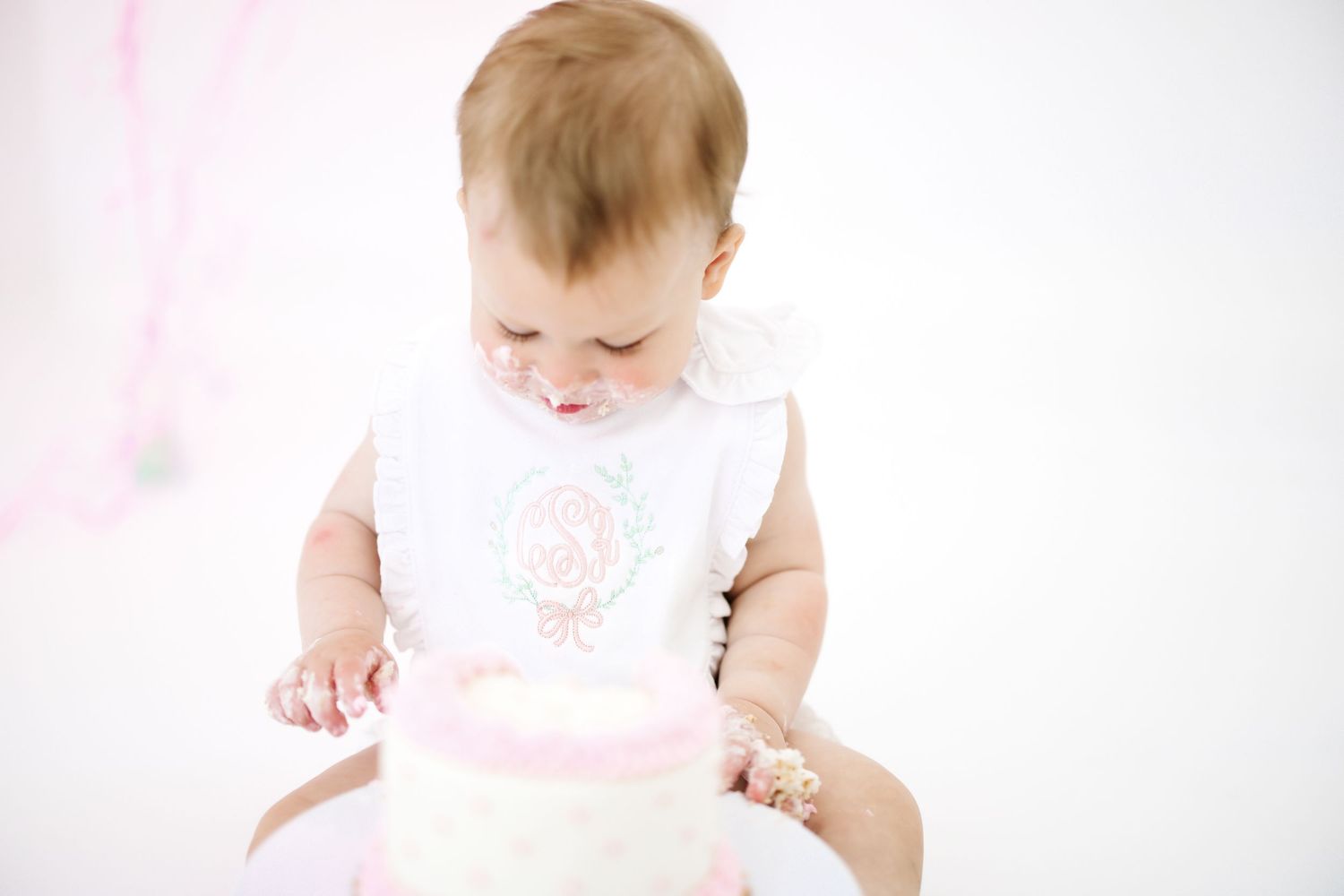 Baby wearing embroidered white dress exploring pink frosted cake.