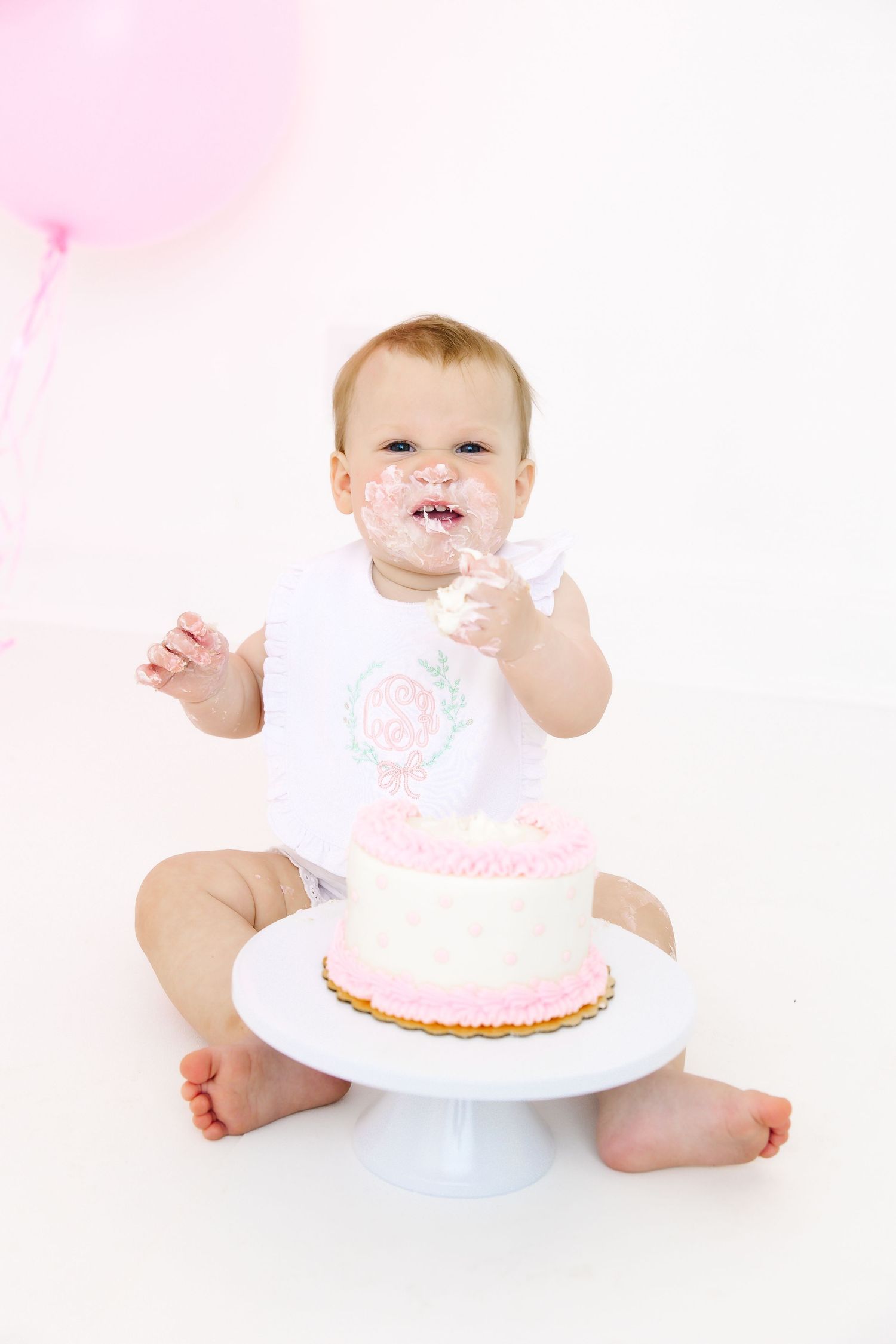 Baby sits on white surface with pink and white birthday cake for a smash cake photo session.
