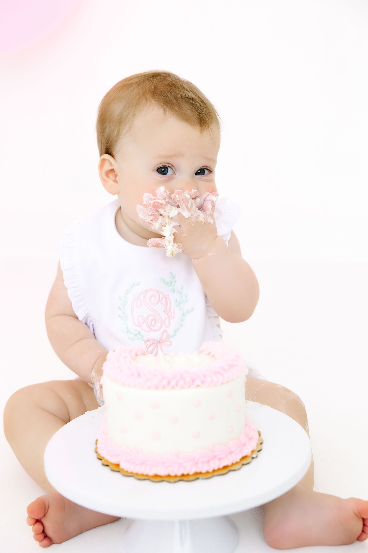 Baby in white outfit enjoying first birthday cake smash with pink frosted cake on white plate.