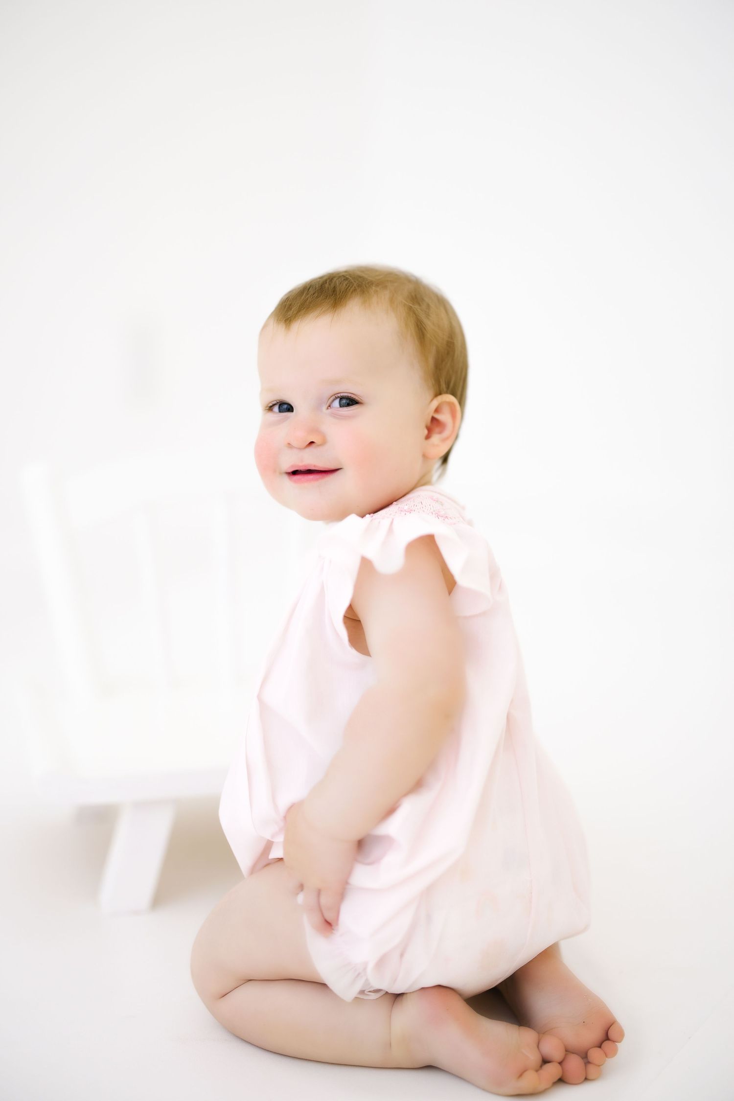 Baby in soft pink romper sitting on white background smiling sweetly during portrait session.
