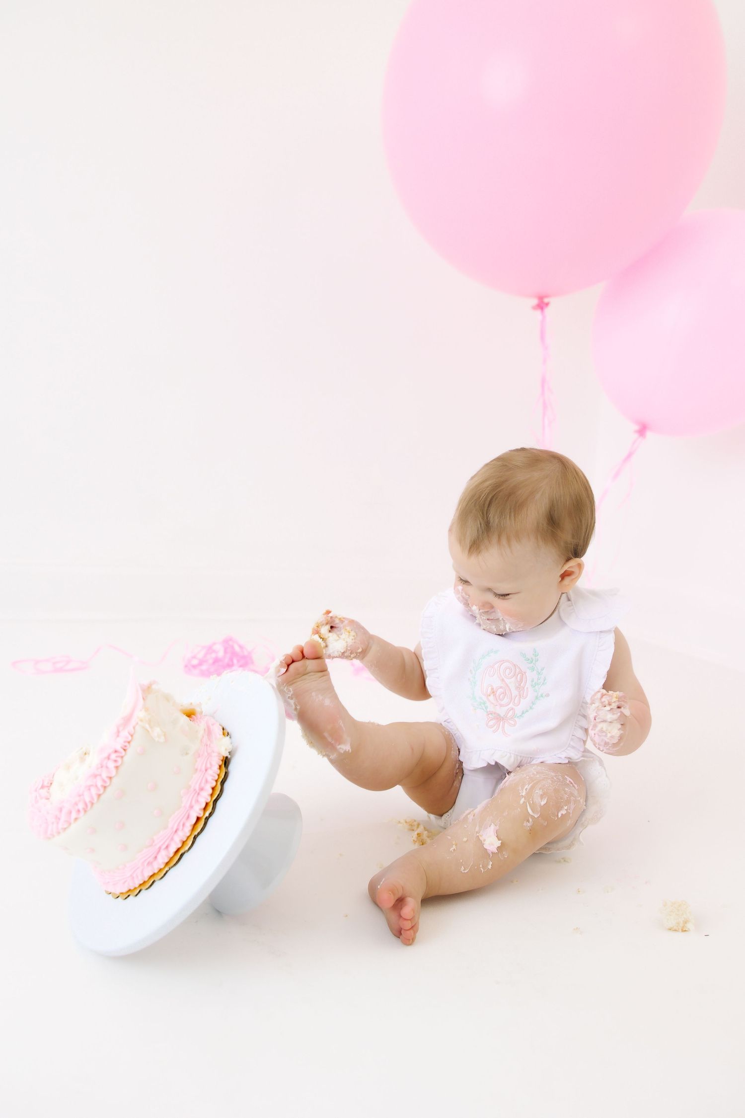 Baby explores first birthday cake with pink balloons in background.