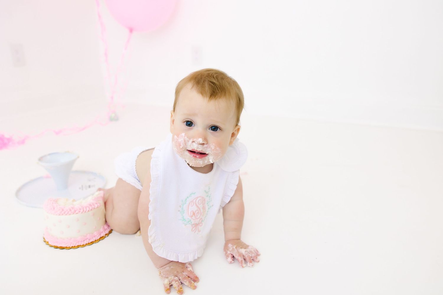 Baby in white bib sits on floor next to pink birthday cake and balloons during first birthday photo session.