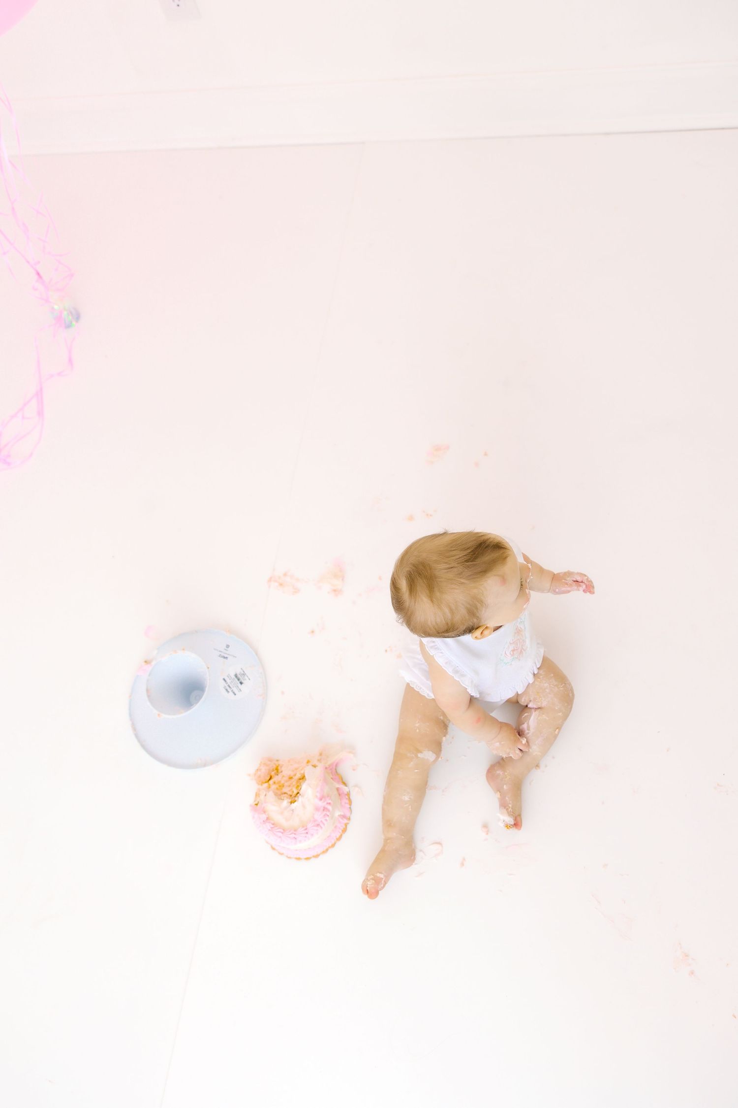 Overhead view of baby playing with smashed cake on white floor with pink accents.
