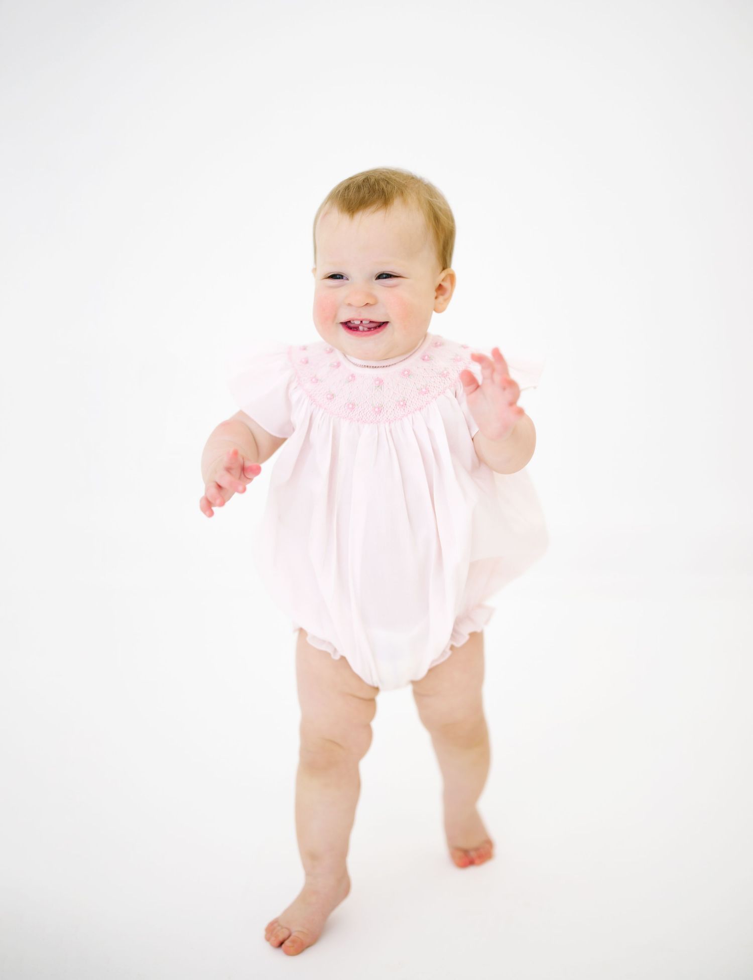Baby wearing pink ruffled outfit smiles while standing on white backdrop.