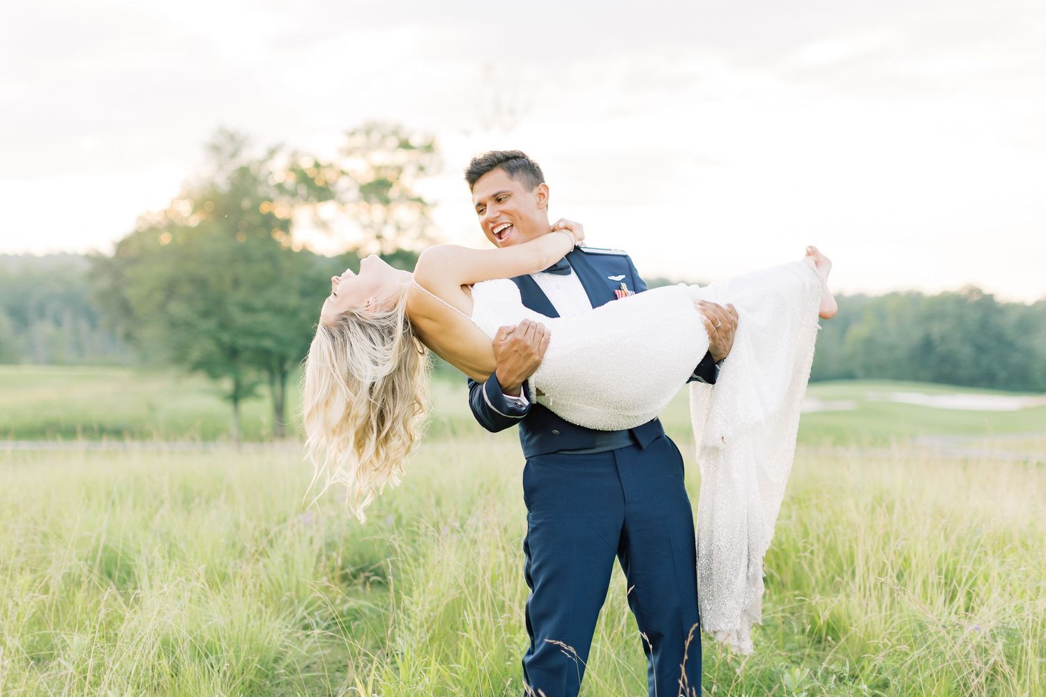 A couple in wedding attire embrace in a sunlit meadow with the groom carrying the bride in a flowing white dress.