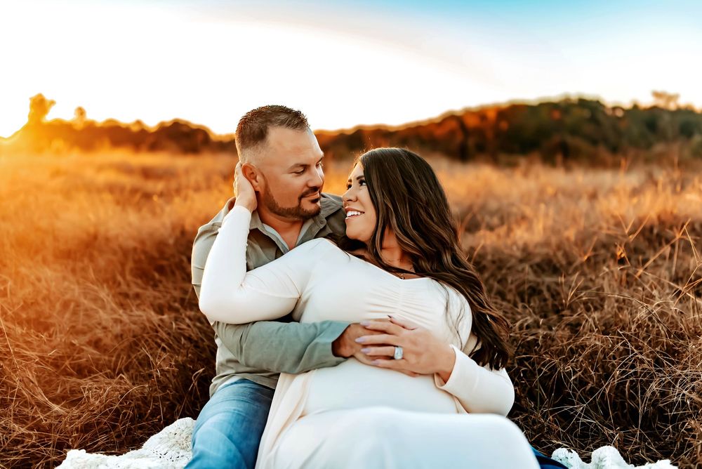 A couple embraces in a sunny field at golden hour with warm lighting and dried grass surrounding them.
