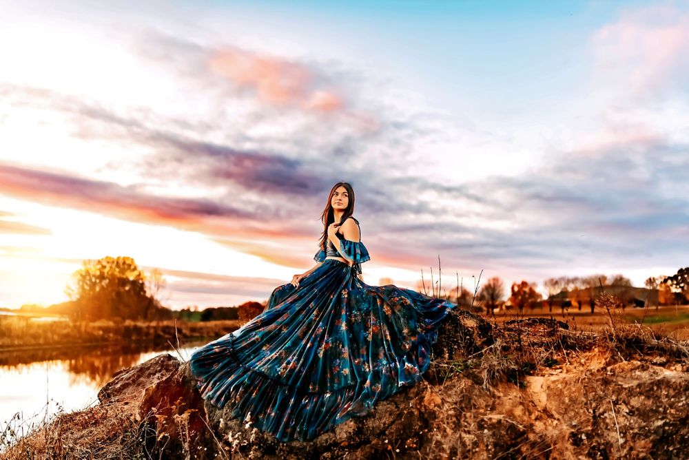 A flowing teal gown sparkles against a stunning sunset landscape with a lake and autumn trees in the background.
