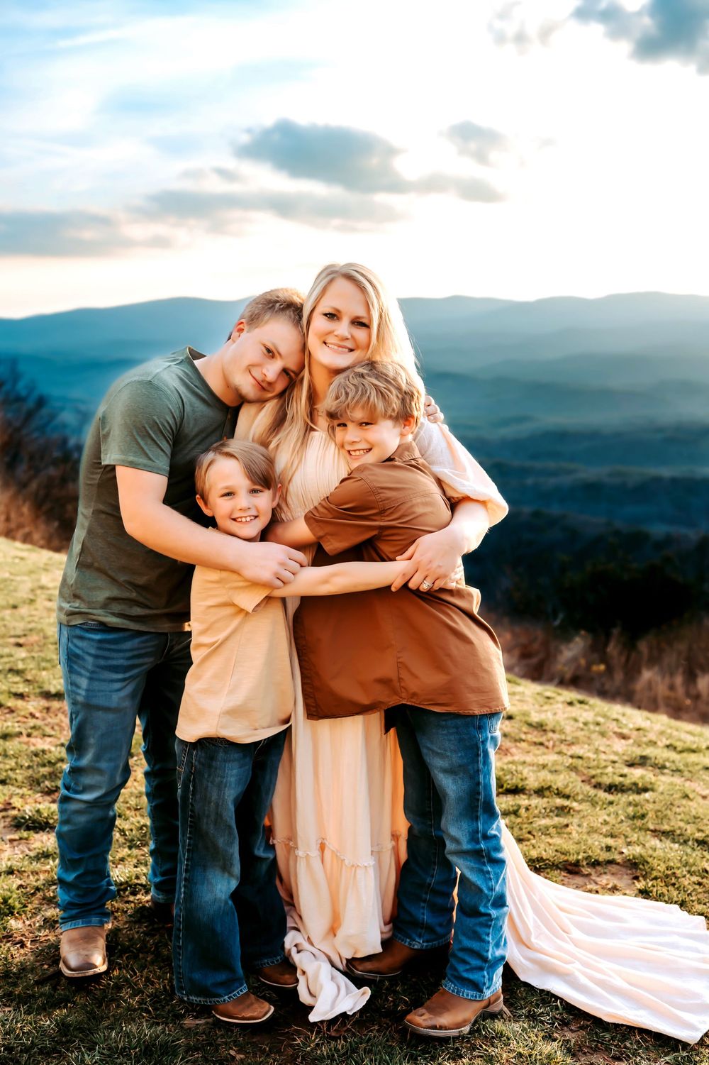 A family embraces together on a grassy hilltop overlooking misty mountains during sunset.