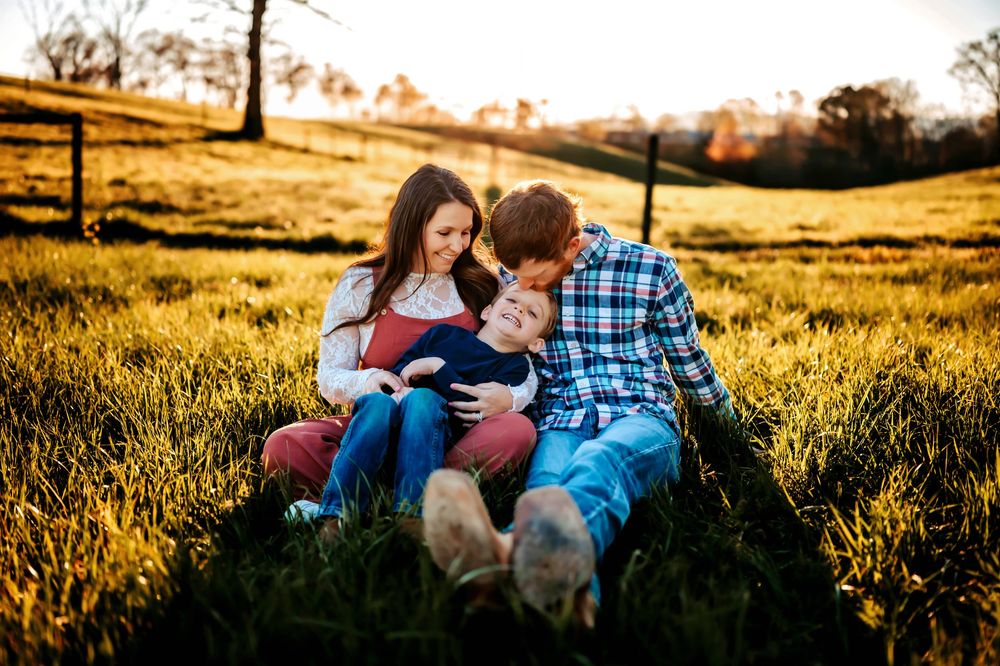 A young family sits together in a sunlit golden field, parents lovingly gazing at their toddler child.