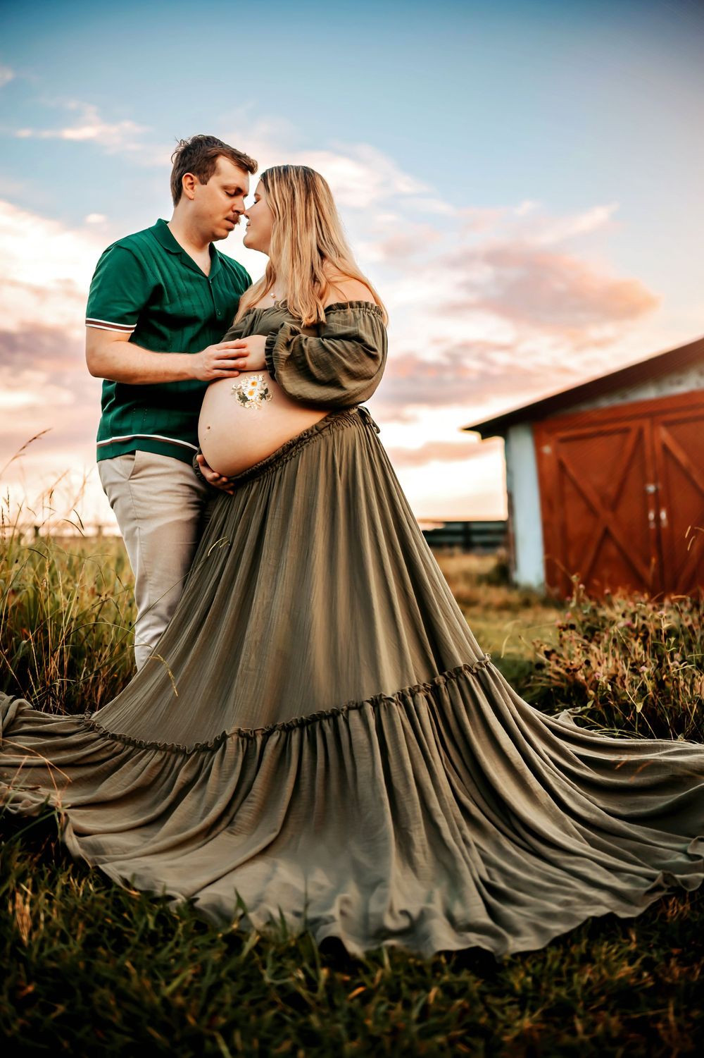 Couple sharing a romantic moment outdoors at sunset, pregnant woman in flowing olive gown with barn backdrop.
