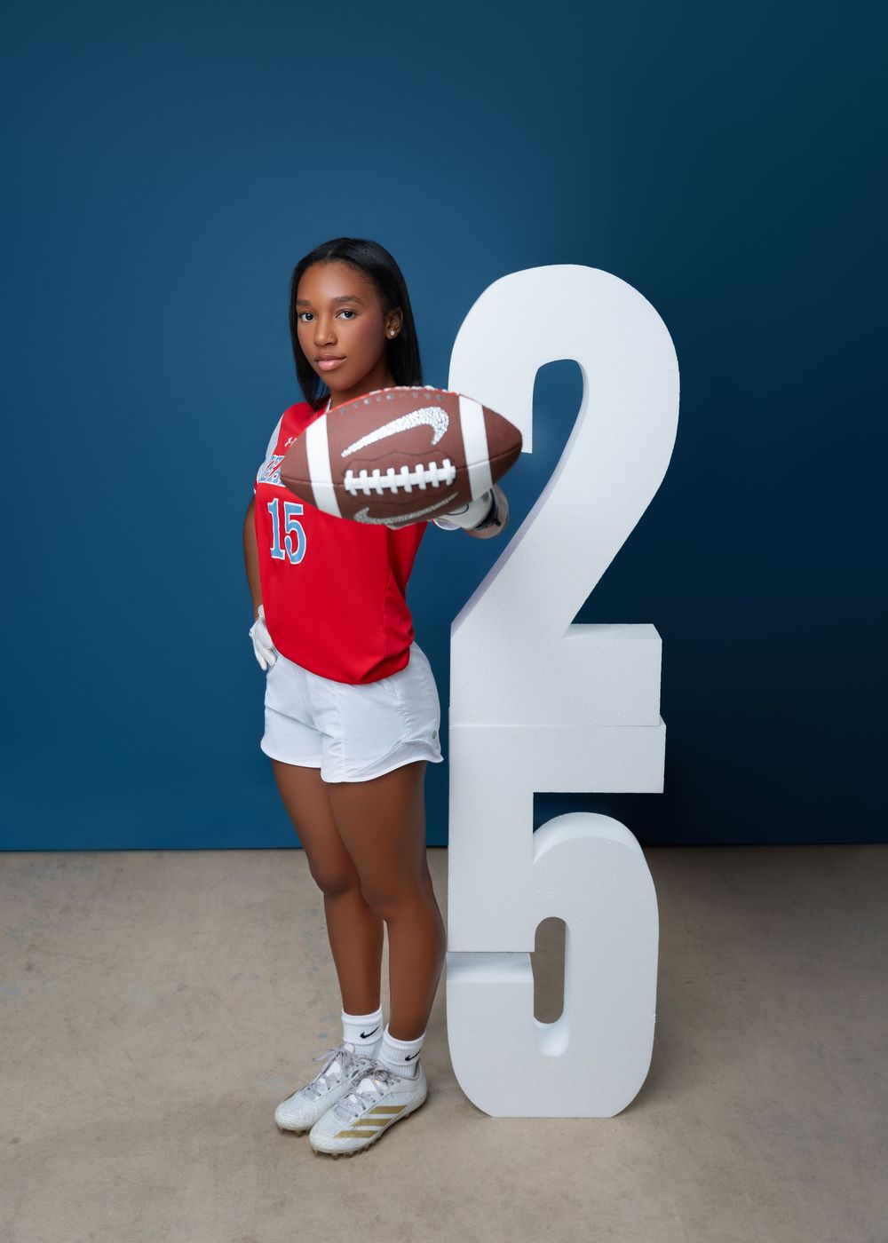 High school athlete in red jersey number 15 poses with football in front of large white number 26 on blue background.