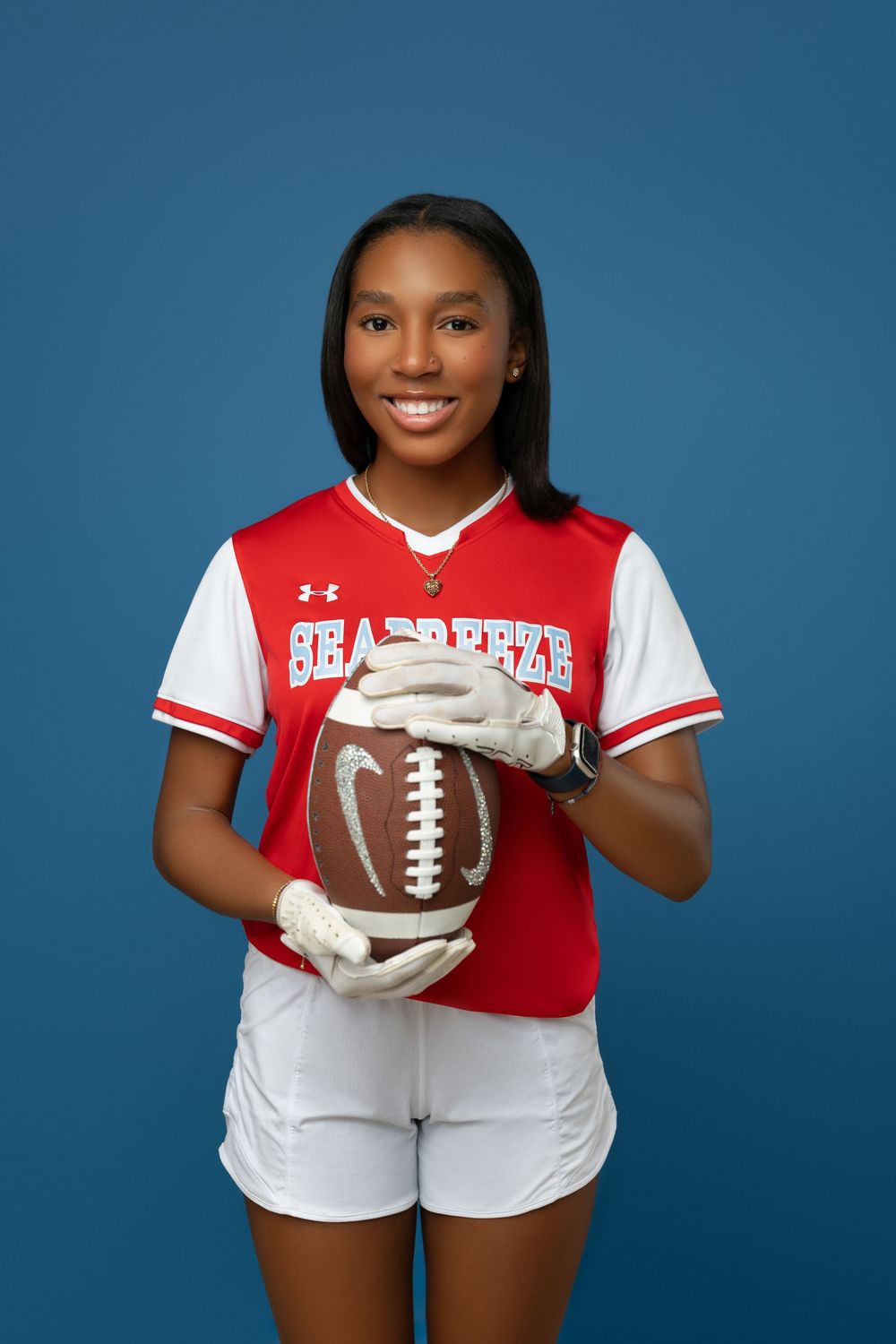 Athletic portrait with football player wearing red and white Under Armour jersey holding football against blue backdrop.