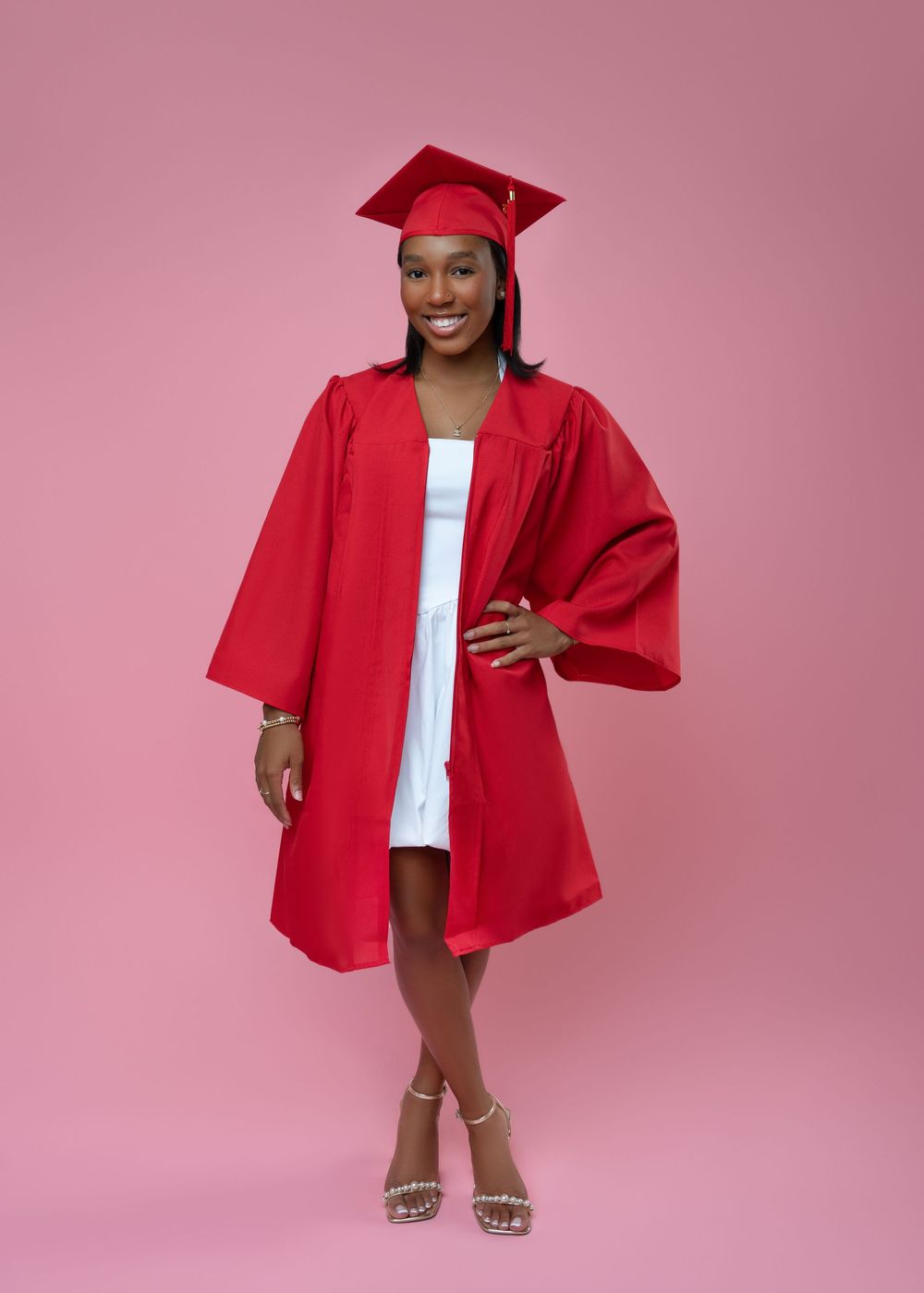 Graduate in bright red cap and gown poses confidently against pink background wearing white dress and sparkly heels.