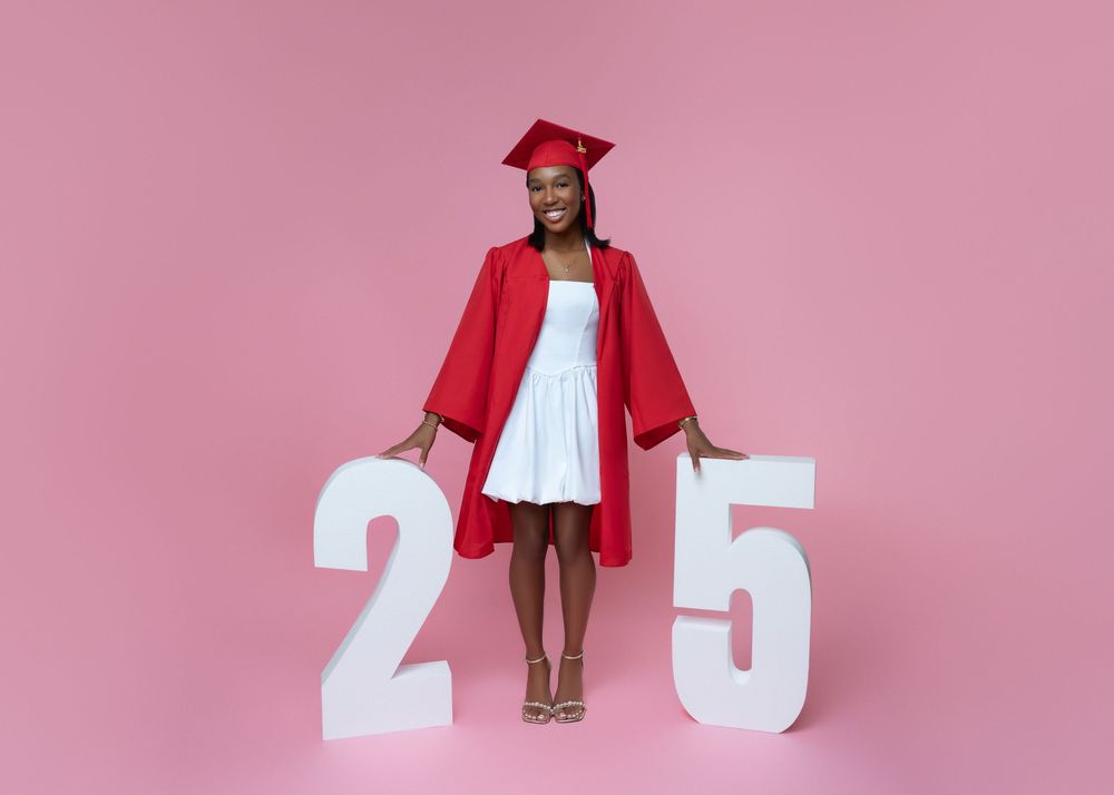 A person in a red graduation gown and cap stands proudly holding a diploma against a pink background.